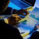 Two businesspeople analyze charts and graphs over a desk with a laptop in the background.