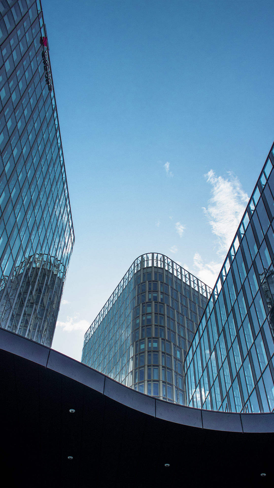 Curved glass office buildings surrounding a blue sky, viewed from street level below.