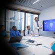 Doctor in a white coat presents brain scan data on two large blue monitors to a team in a conference room.