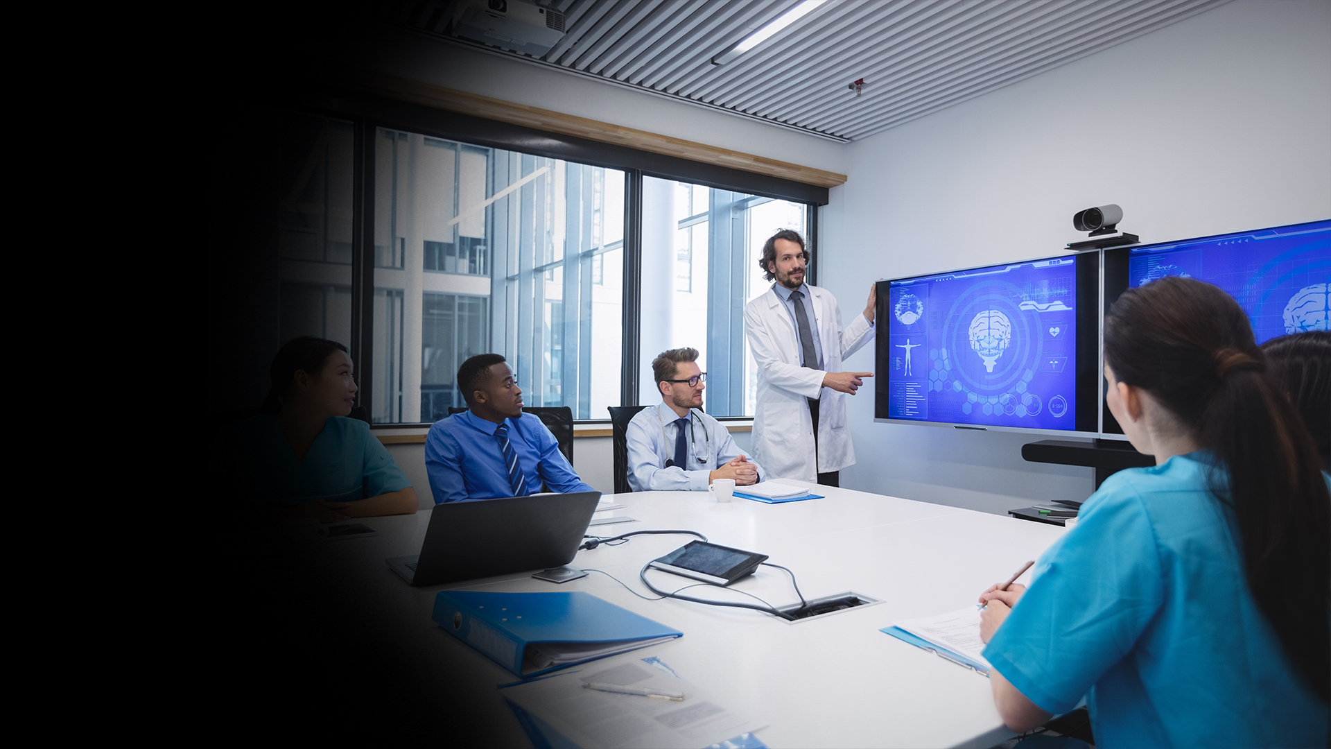 Doctor in a white coat presents brain scan data on two large blue monitors to a team in a conference room.