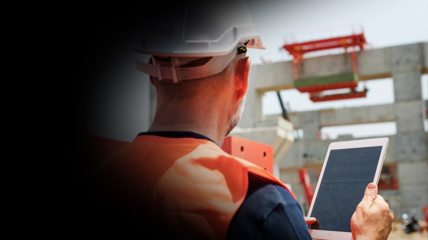 Construction worker in a hard hat and orange safety vest uses a tablet with a checklist at an active building site.