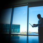 Silhouette of a man in a dress shirt standing by a large window, looking at a tablet in an office setting with a cityscape outside.