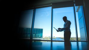 Silhouette of a man in a dress shirt standing by a large window, looking at a tablet in an office setting with a cityscape outside.