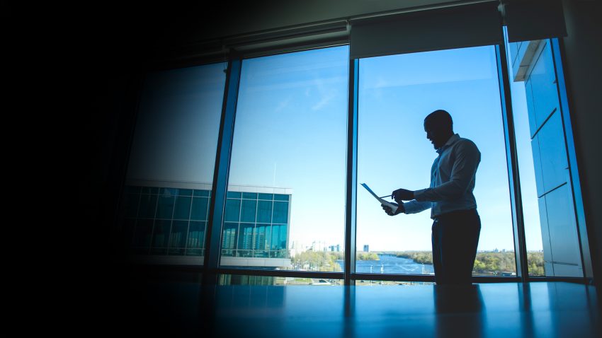 Silhouette of a man in a dress shirt standing by a large window, looking at a tablet in an office setting with a cityscape outside.
