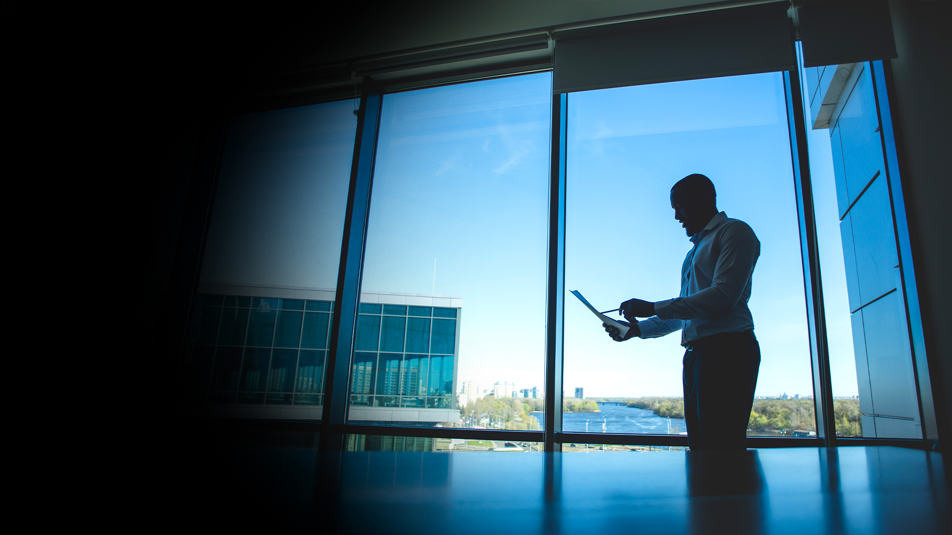 Silhouette of a man in a dress shirt standing by a large window, looking at a tablet in an office setting with a cityscape outside.