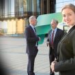 Professional woman in a black suit smiles at the camera outside a glass-office building; two men in suits converse in the background with a green abstract graphic between them.