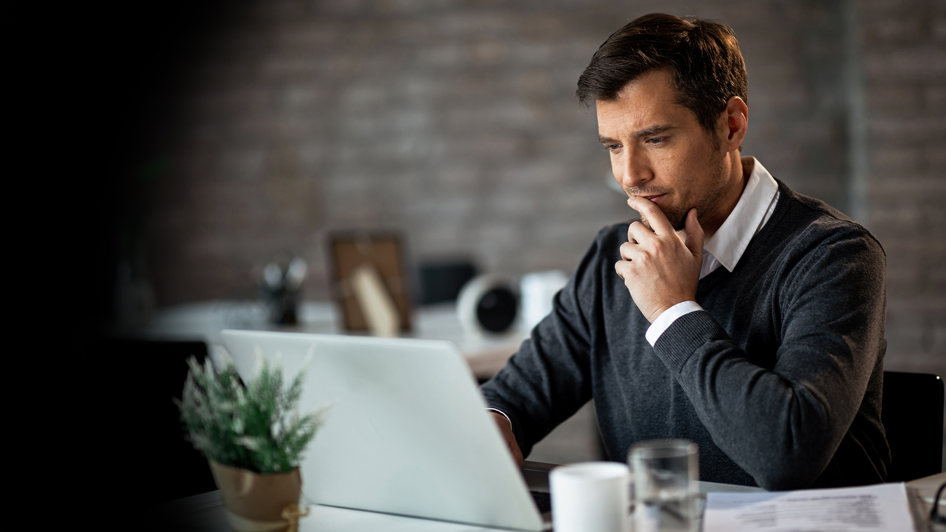 Man sitting at a desk in an office, looking at a laptop and thoughtfully touching his chin, suggesting concentration.