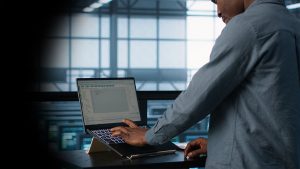 Person standing at a desk, typing on a laptop with a lighted keyboard in a dim control room or data center setting.